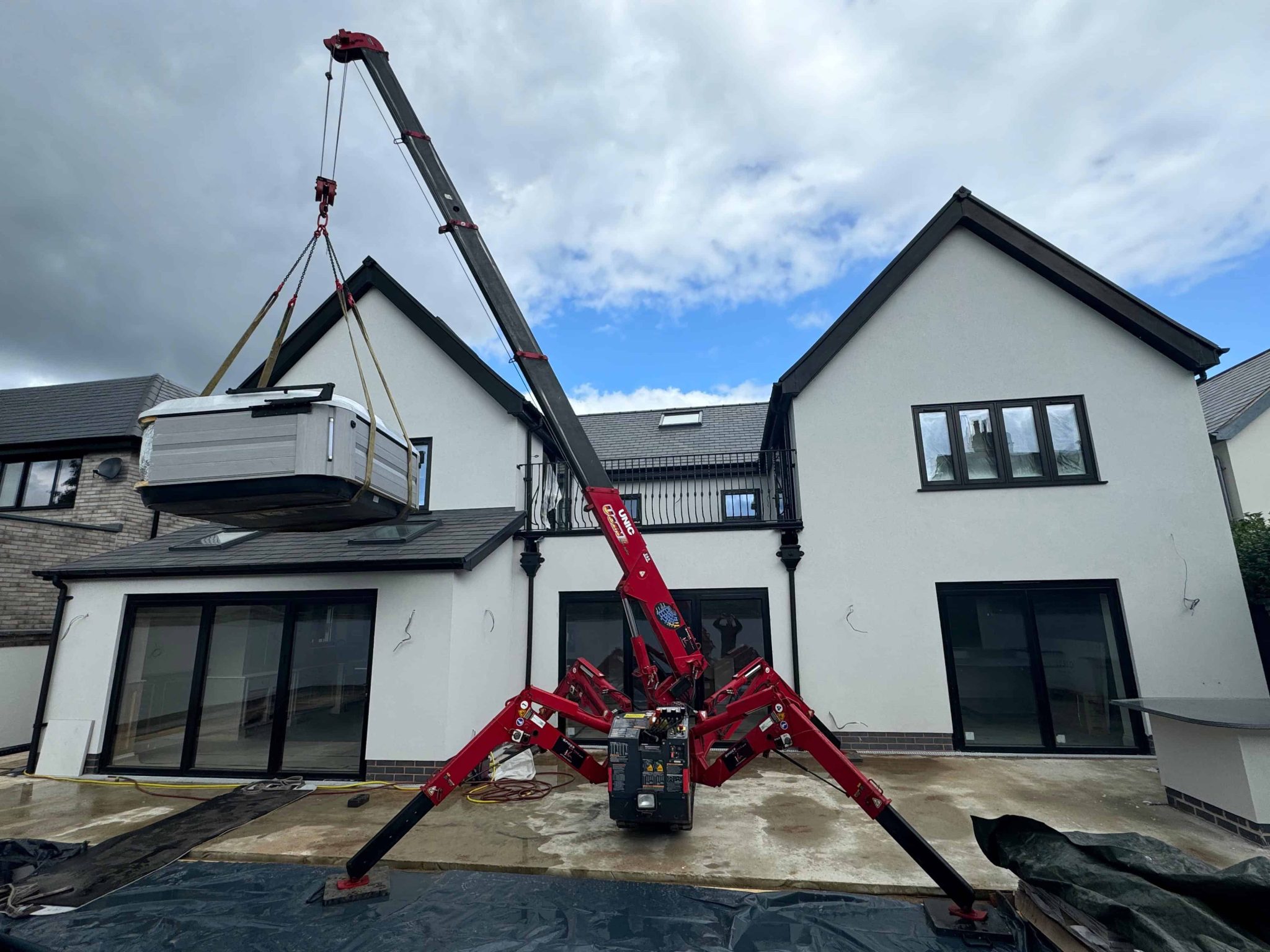 Aerial spider crane lifting a portable hot tub onto the upper terrace of a modern white house during construction or renovation. The spider crane's compact design allows precise lifting in tight spaces.